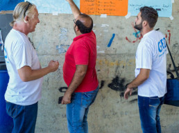 Cap Anamur Team (von links: Volker Rath (Logistiker), Samer Abboud (Dolmetscher) und Addullah Nimje (Krankenpfleger) im Gespräch vor den Plakaten mit den Öffnungszeiten der medizinischen Einrichtungen in Sidon.