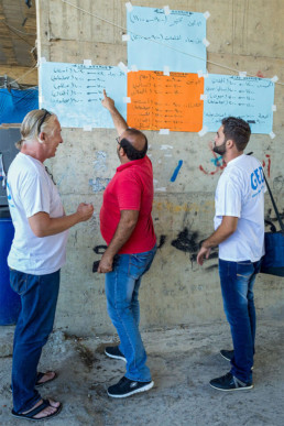 Cap Anamur team (from left: Volker Rath (logistician), Samer Abboud (interpreter) and Addullah Nimje (nurse) talking in front of the posters with the opening hours of the medical facilities in Sidon.
